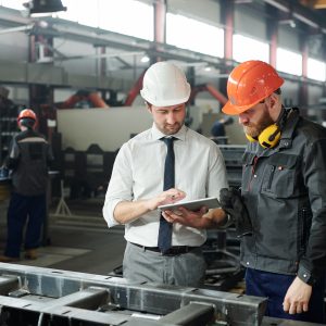 Young master in hardhat and bearded engineer discussing technical sketch on display of tablet in factory workshop